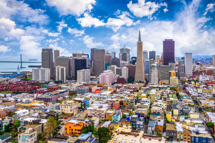 The San Francisco skyline, with multi-colored homes in the foreground before the tall skyscrapers, the sky is blue, and the bay can be seen in the left-hand corner.
