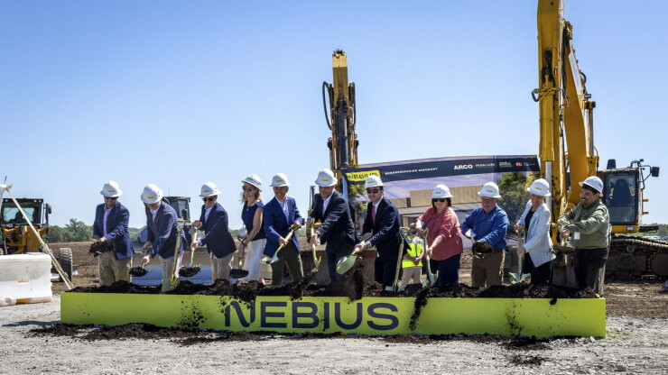 Eleven people in hard hats use shovels to lift dirt over a sign marked 'Nebius'