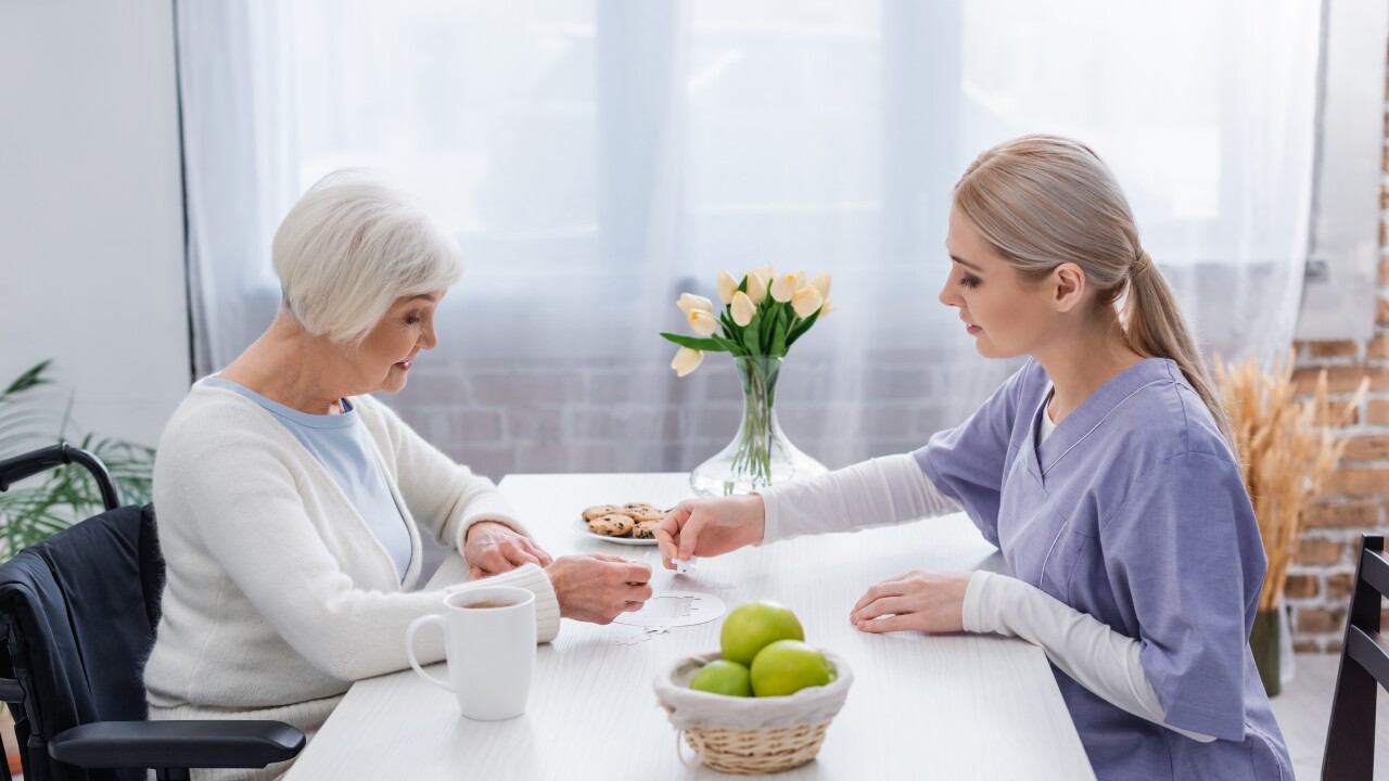 And older woman sits at a table in her home, playing a card game with a female nurse in purple scrubs.