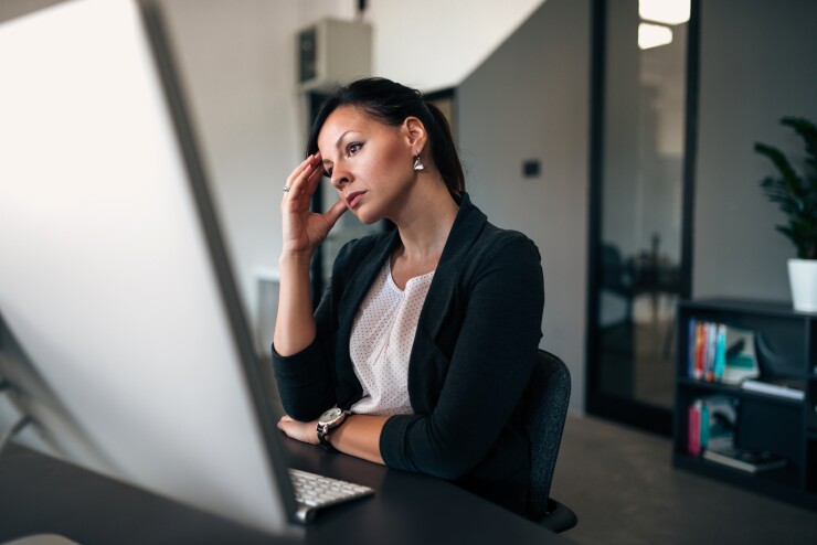 A woman with her hand on her temple looks at her desktop computer in frustration.