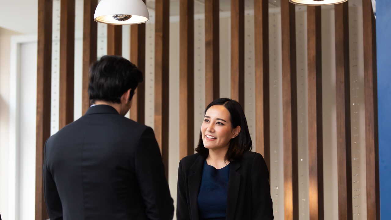 A hotel front desk employee, a woman in a blue top and black blazer, is talking to a man in a black blazer.