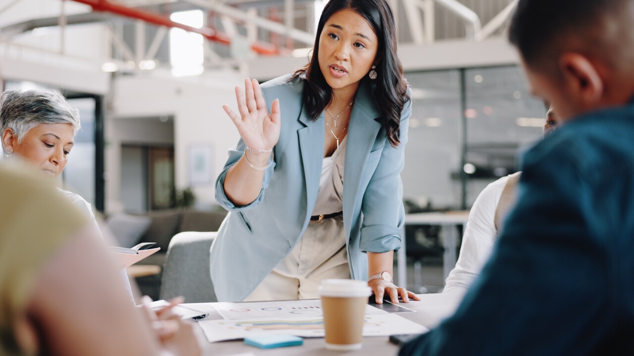 An Asian woman with a light blue blazer stands over a conference table, addressing four colleagues.
