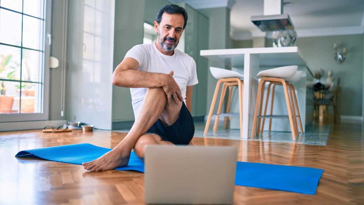 Man sitting on yoga mat stretching looking at laptop computer
