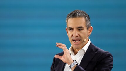 A man in a navy blazer and white shirt speaks with hand gestures and a headset microphone against a blue background.