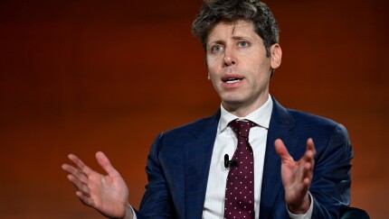 Sam Altman gesturing with open hands while speaking, wearing a blue checkered suit and a maroon polka-dot tie.