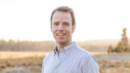 A smiling man in a light grey button-down shirt outdoors in golden hour sunlight with a blurred landscape background.