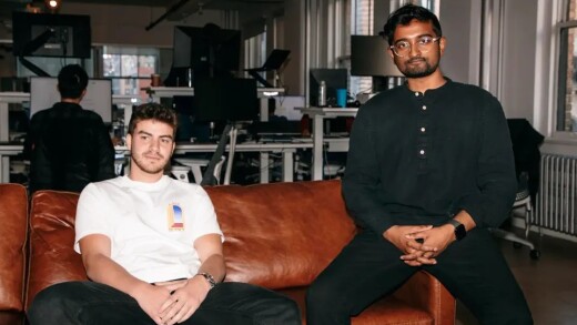 Two men pose in a modern office; one sits on a brown leather sofa, the other perches on the armrest against a desk backdrop.