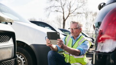 Loss adjuster wearing hi-vis safety vest standing in compound for damaged cars taking photo for insurance claim report on digital tablet