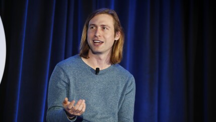 Man with long blond hair and blue tee speaks on a stage with blue curtains in the background.