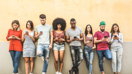 A group of young people against a wall, looking at their smart phones.