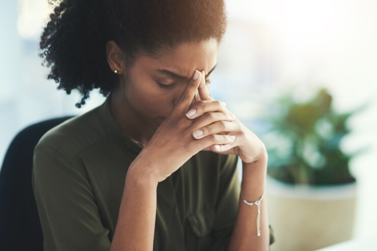 A young Black woman has her head down at her desk, looking exhausted.