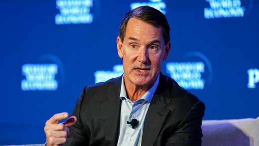 A man in a dark suit speaks at a panel, gesturing with his hand in front of a blue background.