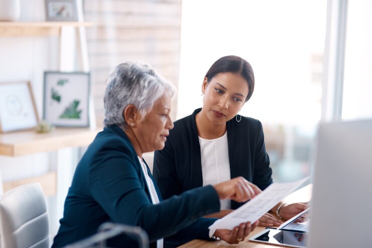 A elderly woman is explaining something to a younger woman; they are in a sunlit office.