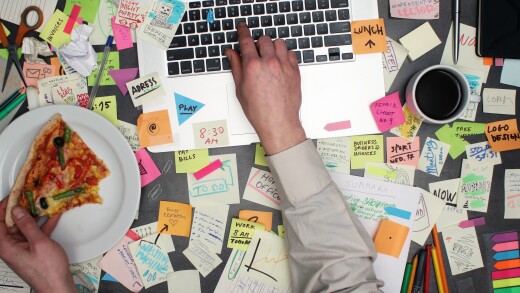 A person eats pizza while working at a laptop surrounded by to-do notes.