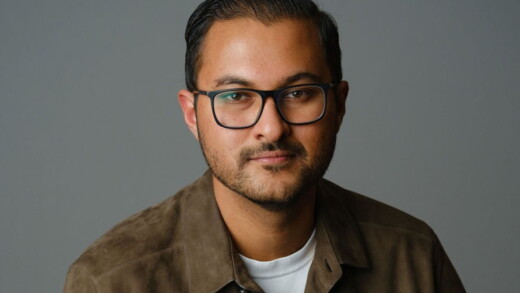 Headshot of a man with black glasses and short beard, wearing a brown suede shirt against a solid gray background.