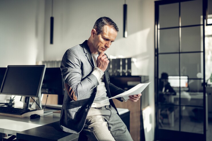 Man in sport jacket at work leaning against desk reading paper thinking