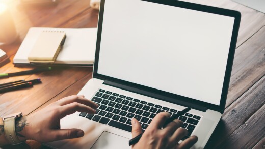 A person typing on a laptop with a pen in one of their hands