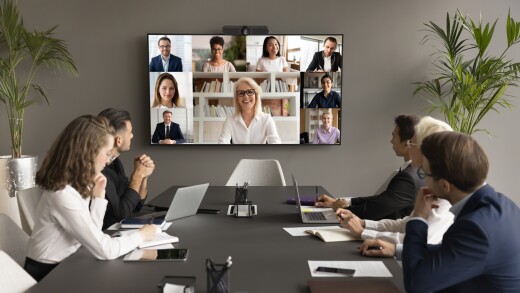 People around a board table speaking with colleagues on a television monito on the wall.