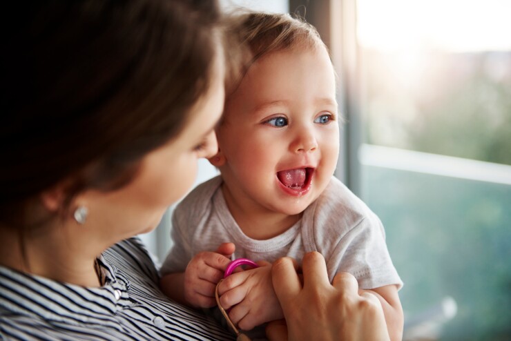 A woman holds her laughing baby; they are next to window with light shining through it.