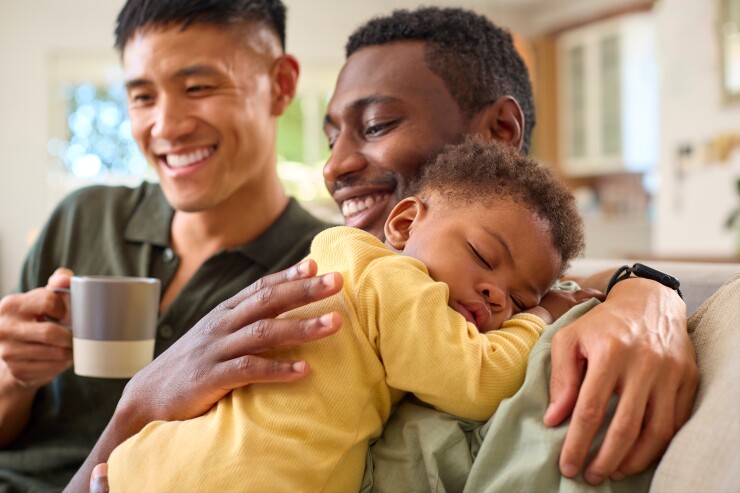 Two dads happily sit together on their couch. One dad holds their baby.
