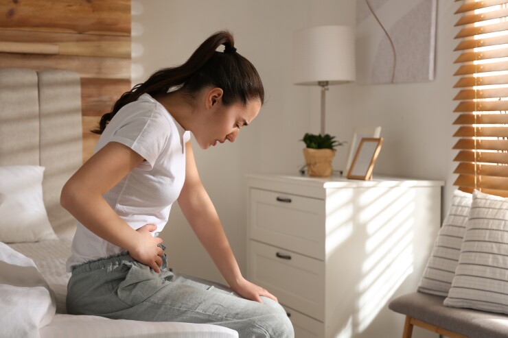 A young woman sits at the edge of her bed, clutching her stomach in pain.