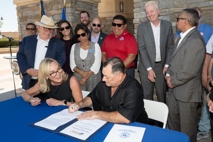 Two people at a table signing documents with at least nine people standing behind them
