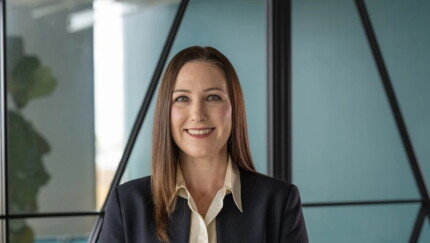 A smiling woman with straight brown hair wearing a dark blazer and cream collared shirt in a modern office setting.