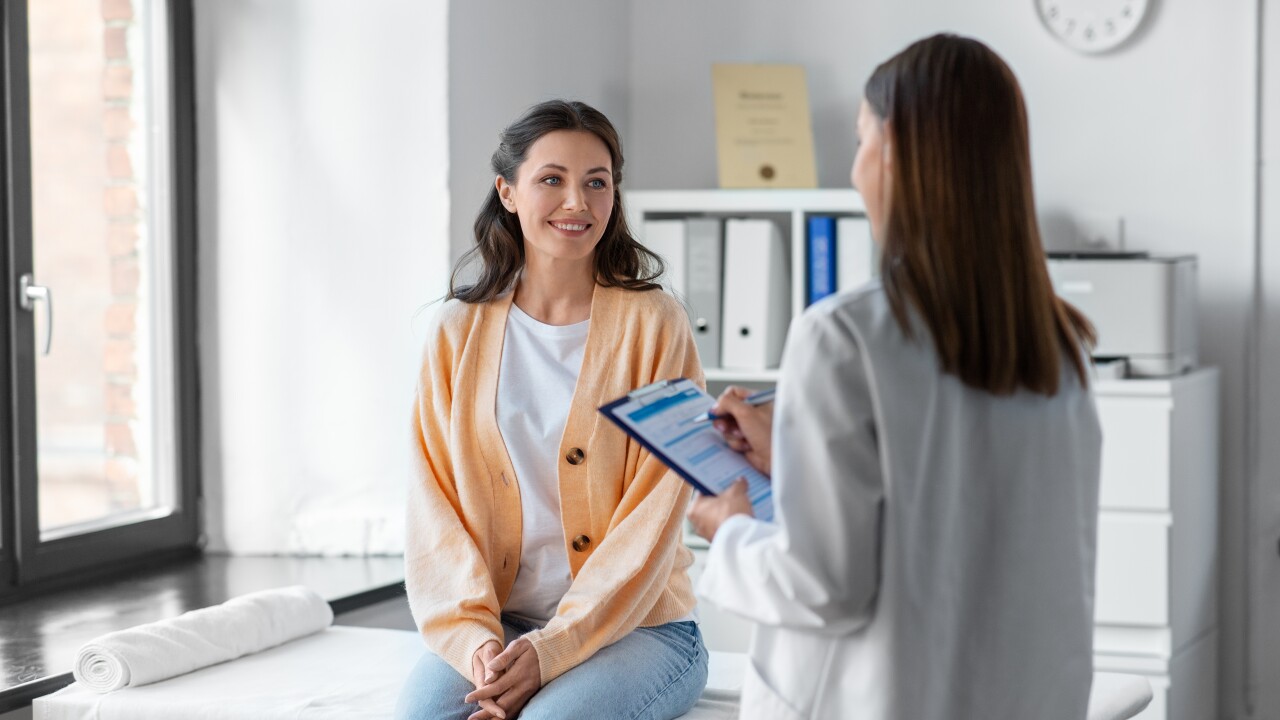A woman in an orange cardigan sits on an examining table while a doctor with a clipboard asks her questions