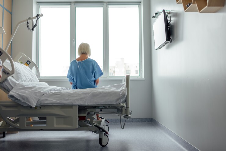 A woman in a blue hospital gown is sitting on a bed alone, staring out the window; her back faces the camera.