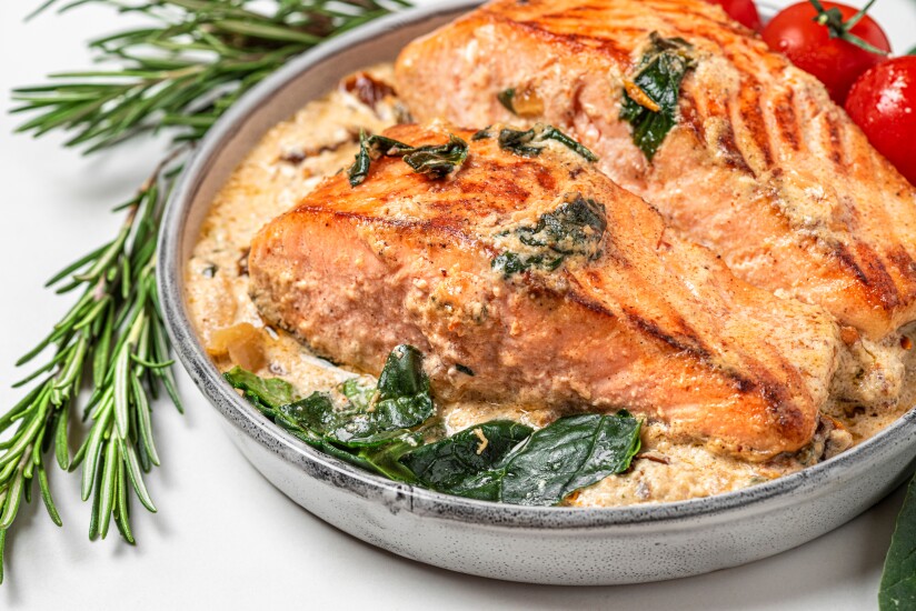 Close-up of grilled salmon steak with cream sauce, spinach, tomatoes and rosemary on a white marble background.