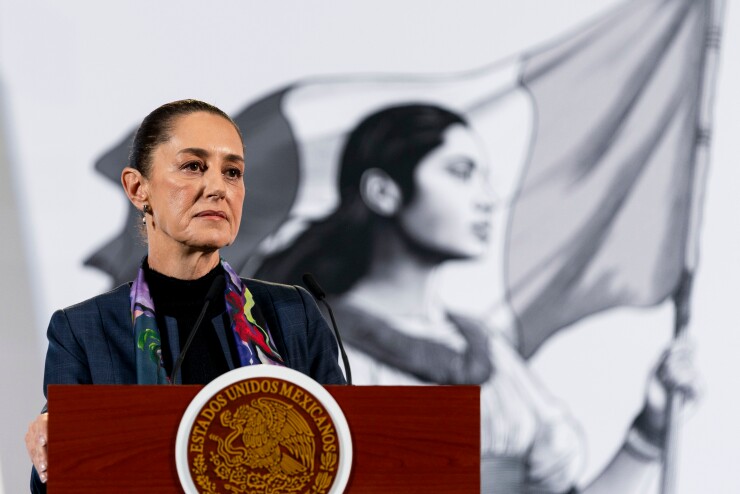 Claudia Sheinbaum posing behind a lectern in front of a painting of a woman holding the Mexican flag