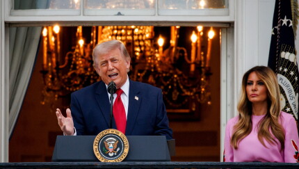 President Trump speaks at an outdoor White House lectern with a lit chandelier in the background, with Melania Trump in a pink dress standing behind him.