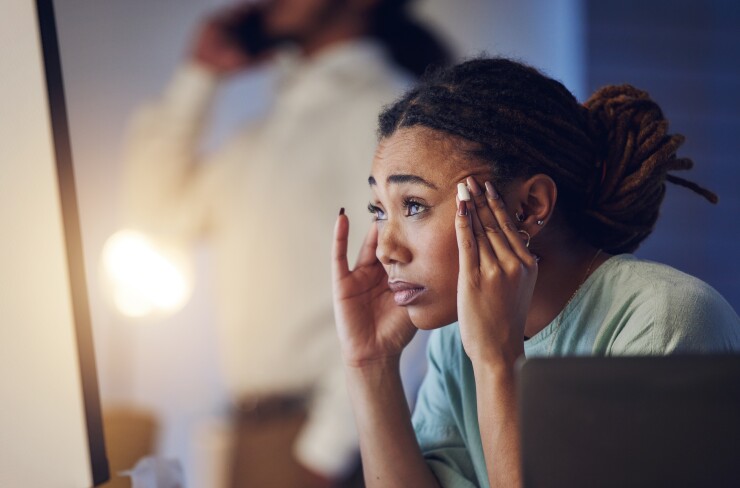 A young Black woman rubs her temples, looking pained; she is turned away from her laptop.