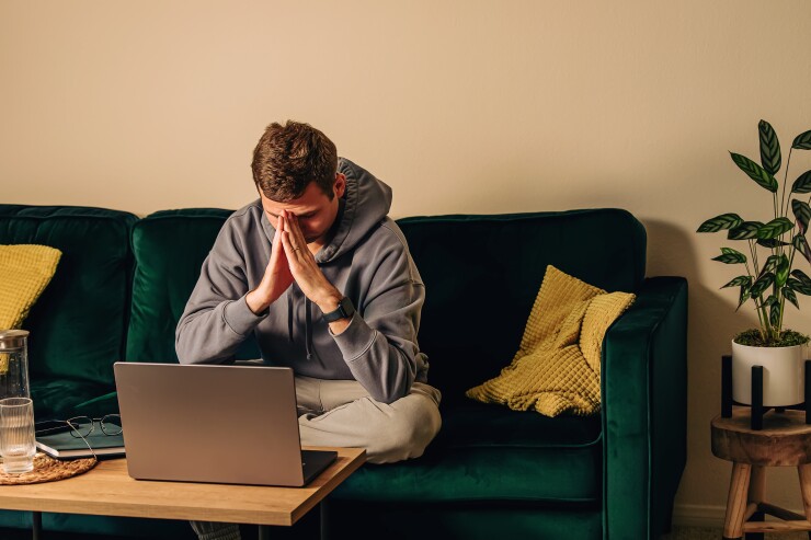 A white man looks down in frustration at his laptop while sitting on a velvet green couch.