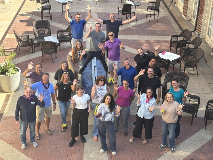 Employees wave from below the camera on a brick sidewalk dotted with chairs and tables.