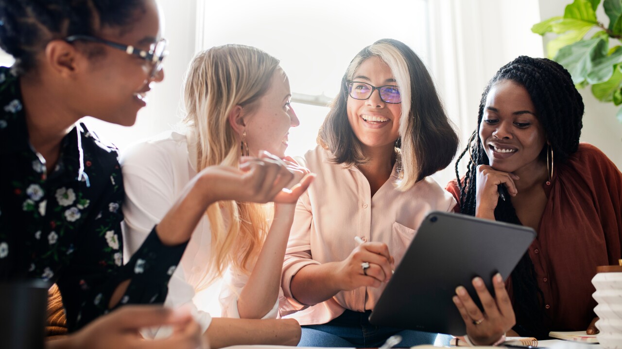 Four women gather around an iPad, smiling; they are backlit by sunlight streaming through a window.
