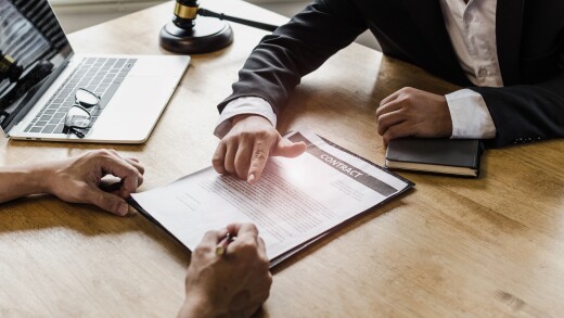 Client signing a contract with a lawyer. There's an open laptop and a wooden judge gavel on the desk.