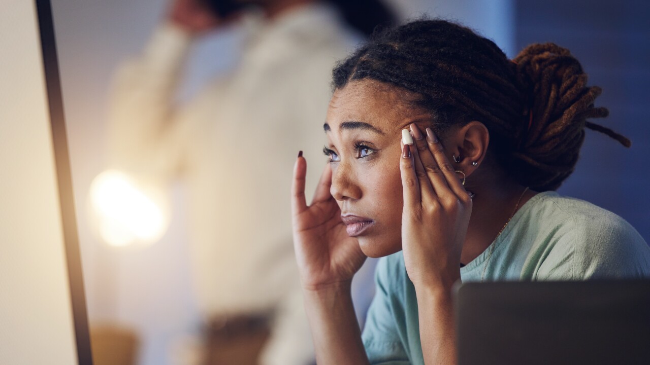 A young Black woman rubs her temples, looking pained; she is turned away from her laptop.
