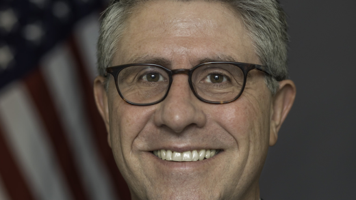 Headshot of man with gray hair and glasses wearing a suit and patterned pink tie standing in front of a blurred American flag in the background.