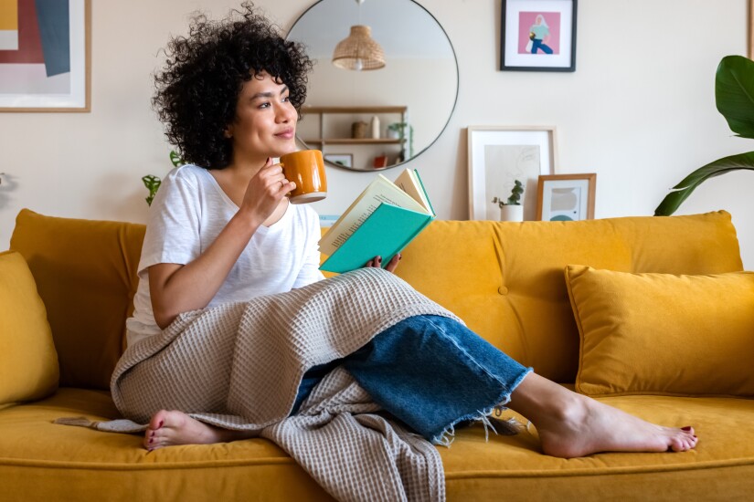 A woman sits on her yellow couch with a cup of coffee and a teal-colored book.