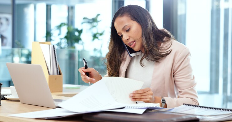 Woman on phone, looking at papers with open laptop on desk