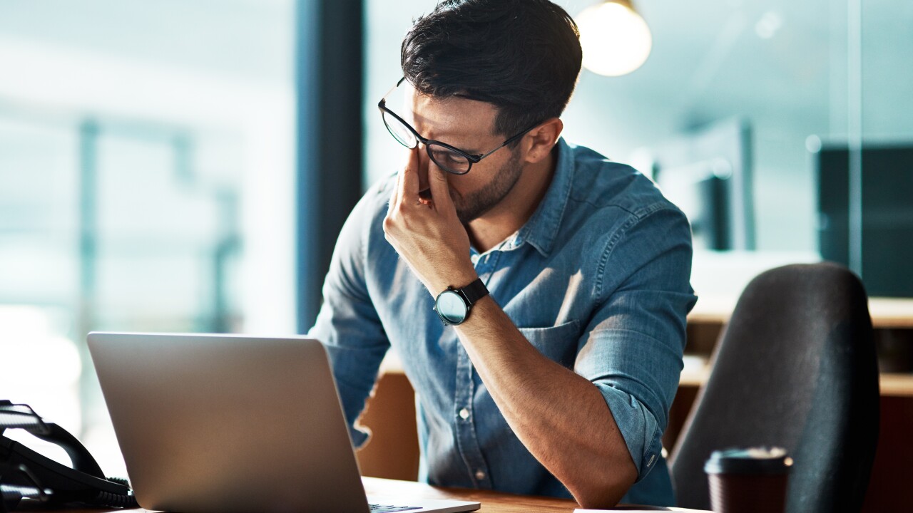 A man with glasses is pinching the bridge of his nose in frustration while sitting in front of his laptop.