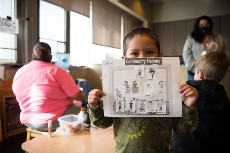 A young child holds up a piece of paper labeled "community helpers," with adults in the background helping other kids.