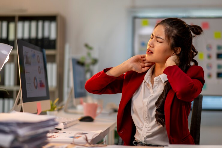 Female employee sitting at desk rubbing back of neck; uncomfortable