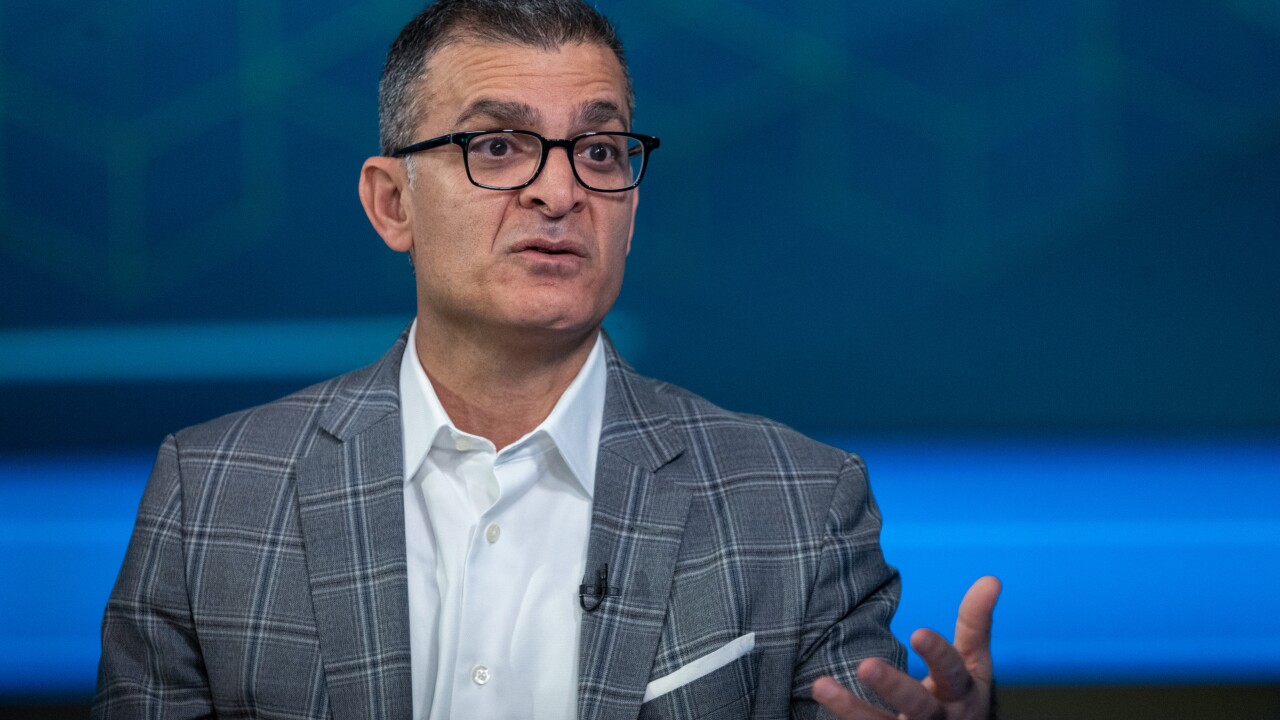 A man with glasses and a grey plaid blazer gestures while speaking against a blue, geometric patterned background.