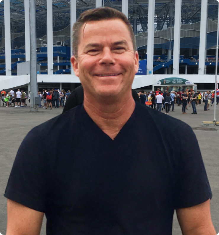 Man with brown hair and black collard shirt stands outside of a stadium entrance.