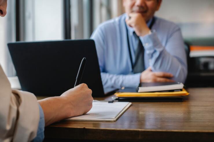 A consultant and a client face each other at a desk in an office; one is taking notes while the other looks on.
