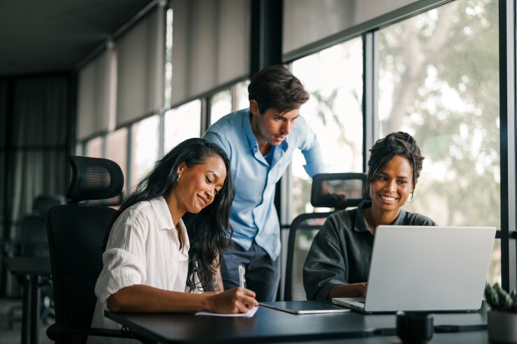 Three employees working together, looking at computer, writing, smiling