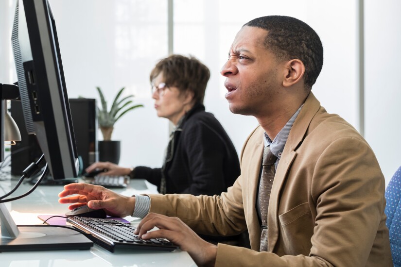 A businessman is staring at his desktop computer in confusion. He is in the office, sitting at a long white table with another colleague next to him.