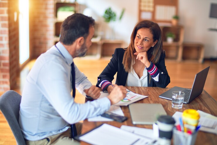 Two employees discussing, sitting at table with laptops and paperwork